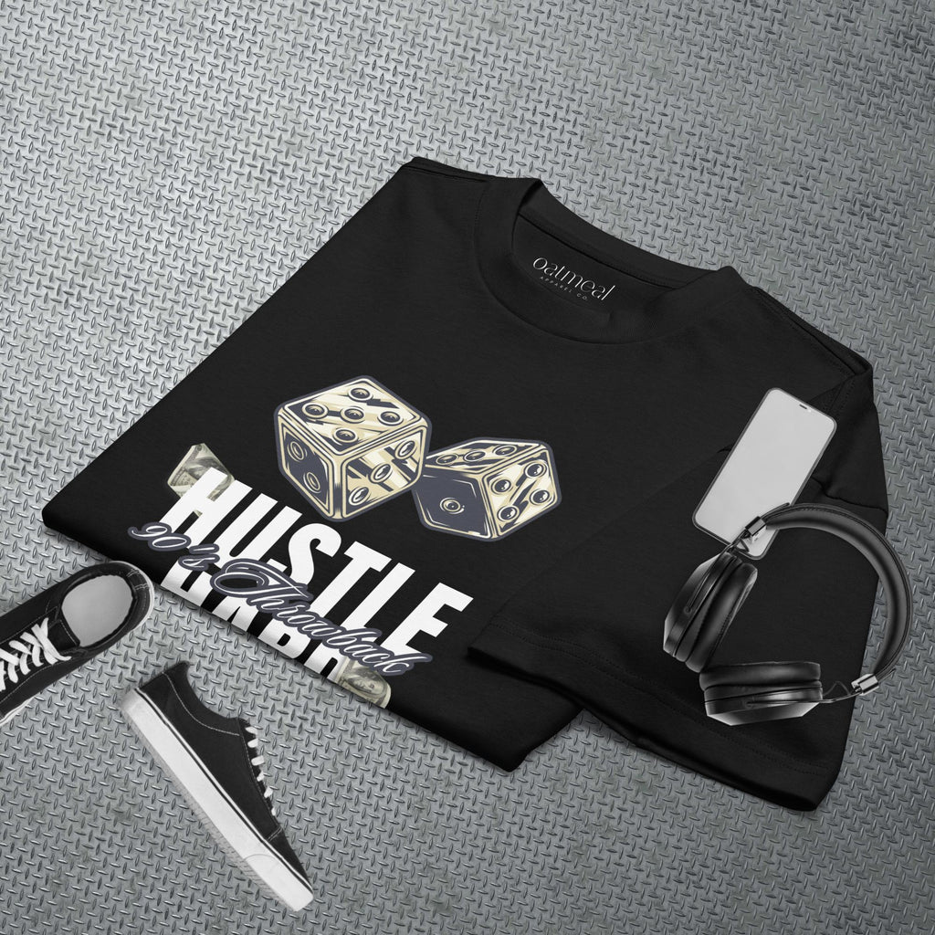 Black t-shirt with dice design and text, placed on a textured gray surface with accessories at Oatmeal Apparel Co.