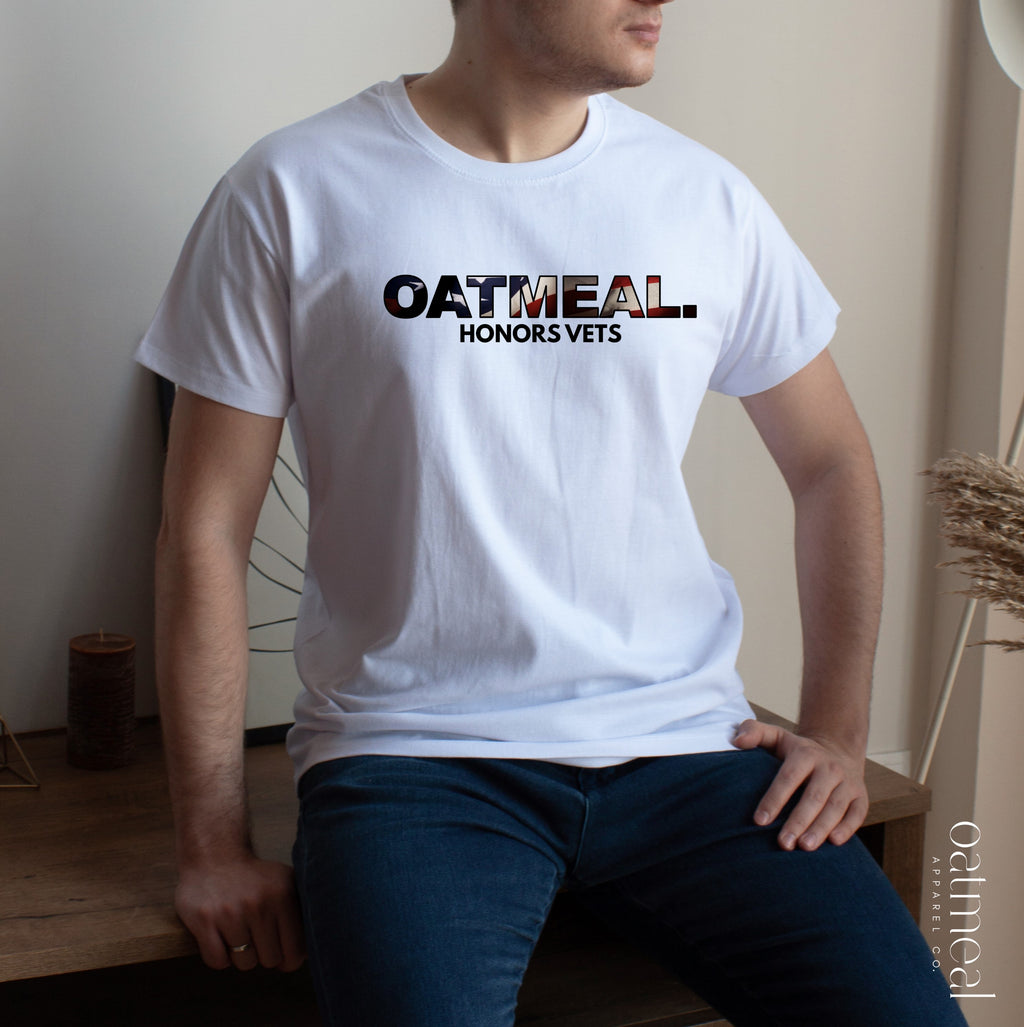 Person wearing a white t-shirt with 'OATMEAL HONORS VETS' text, sitting on a wooden bench at Oatmeal Apparel Co.