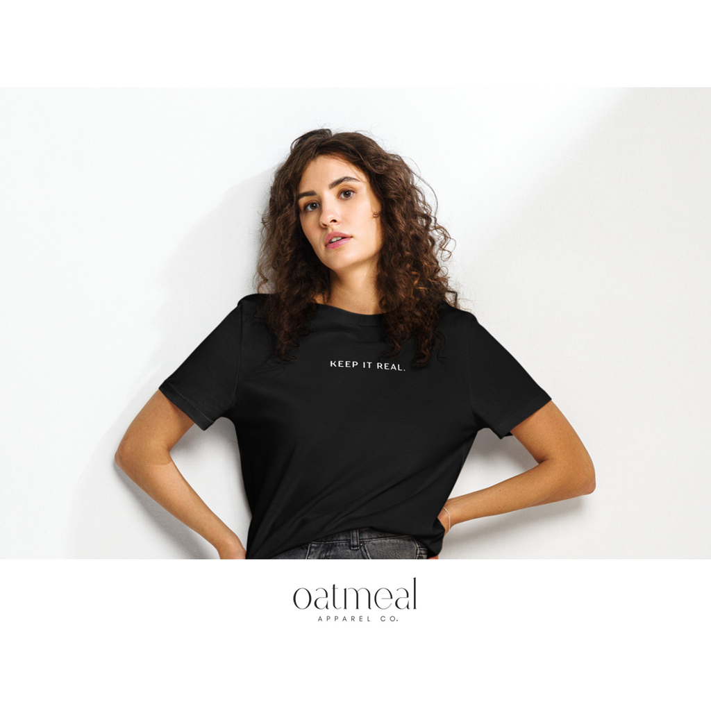 Woman wearing a black t-shirt with 'KEEP IT REAL' text, standing against a white wall with 'oatmeal apparel co.' branding.