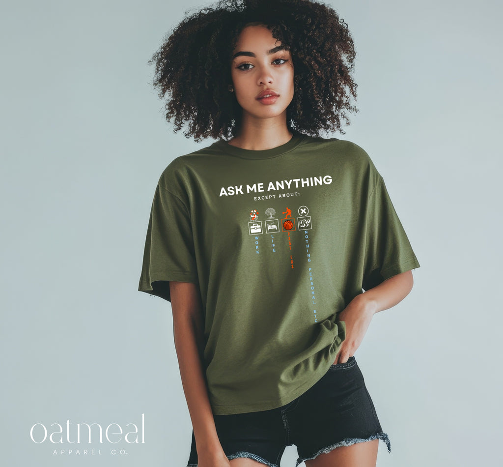A woman modeling an oversized olive green graphic t-shirt with the text 'ASK ME ANYTHING EXCEPT ABOUT' and small illustrated icons, paired with black denim shorts against a light gray studio background. The oatmeal apparel co at Oatmeal Apparel Co.