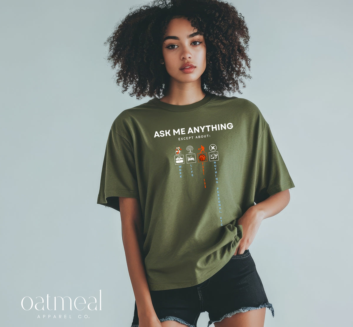 A woman modeling an oversized olive green graphic t-shirt with the text 'ASK ME ANYTHING EXCEPT ABOUT' and small illustrated icons, paired with black denim shorts against a light gray studio background. The oatmeal apparel co at Oatmeal Apparel Co.