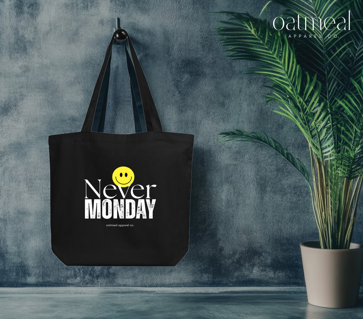 A black canvas tote bag with 'Never MONDAY' and a yellow smiley face graphic hanging on a hook against a textured blue-gray wall, with a potted green plant on the right and the oatmeal apparel logo in the top right corner at Oatmeal Apparel Co.