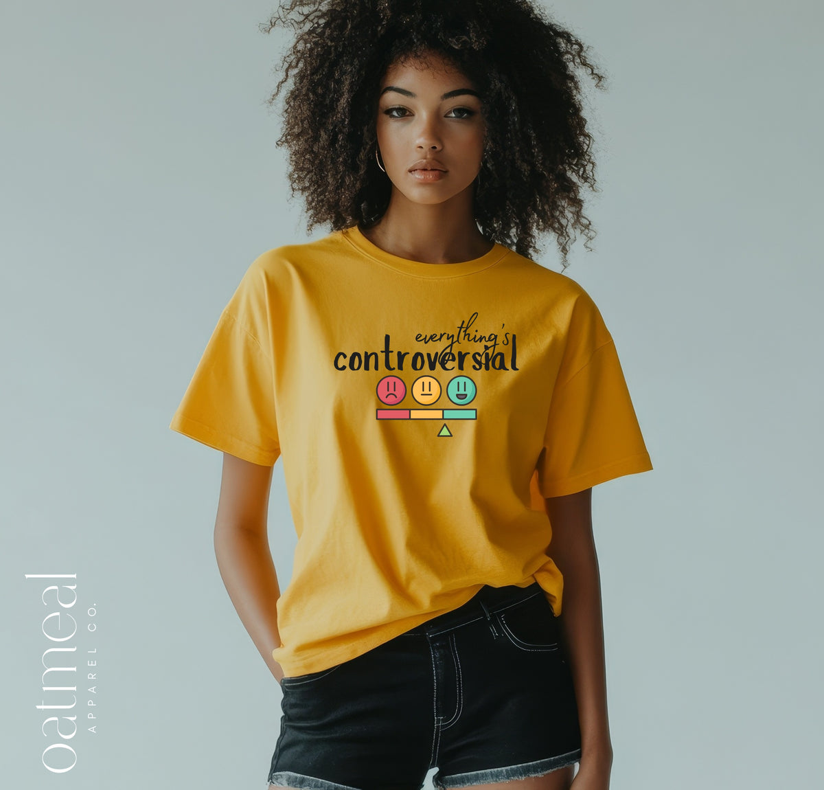 A model with curly dark hair wearing a bright yellow graphic t-shirt that reads 'everything controversial' above a colorful scale graphic, paired with black denim shorts at Oatmeal Apparel Co.