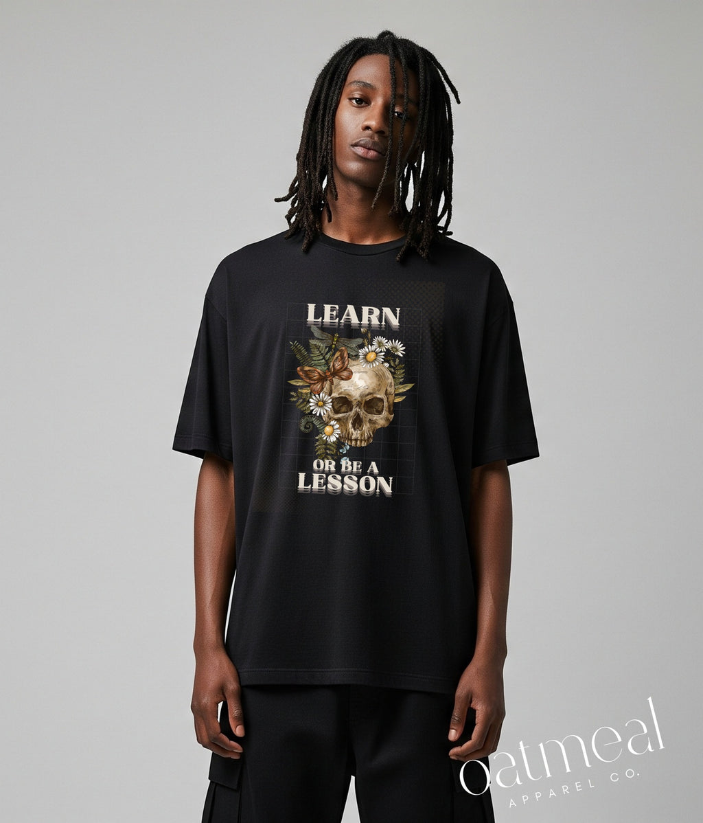 A model with long dreadlocks stands against a light gray studio background wearing a black graphic t-shirt with skull and flowers and the text 'LEARN OR BE A LESSON' design at Oatmeal Apparel Co.