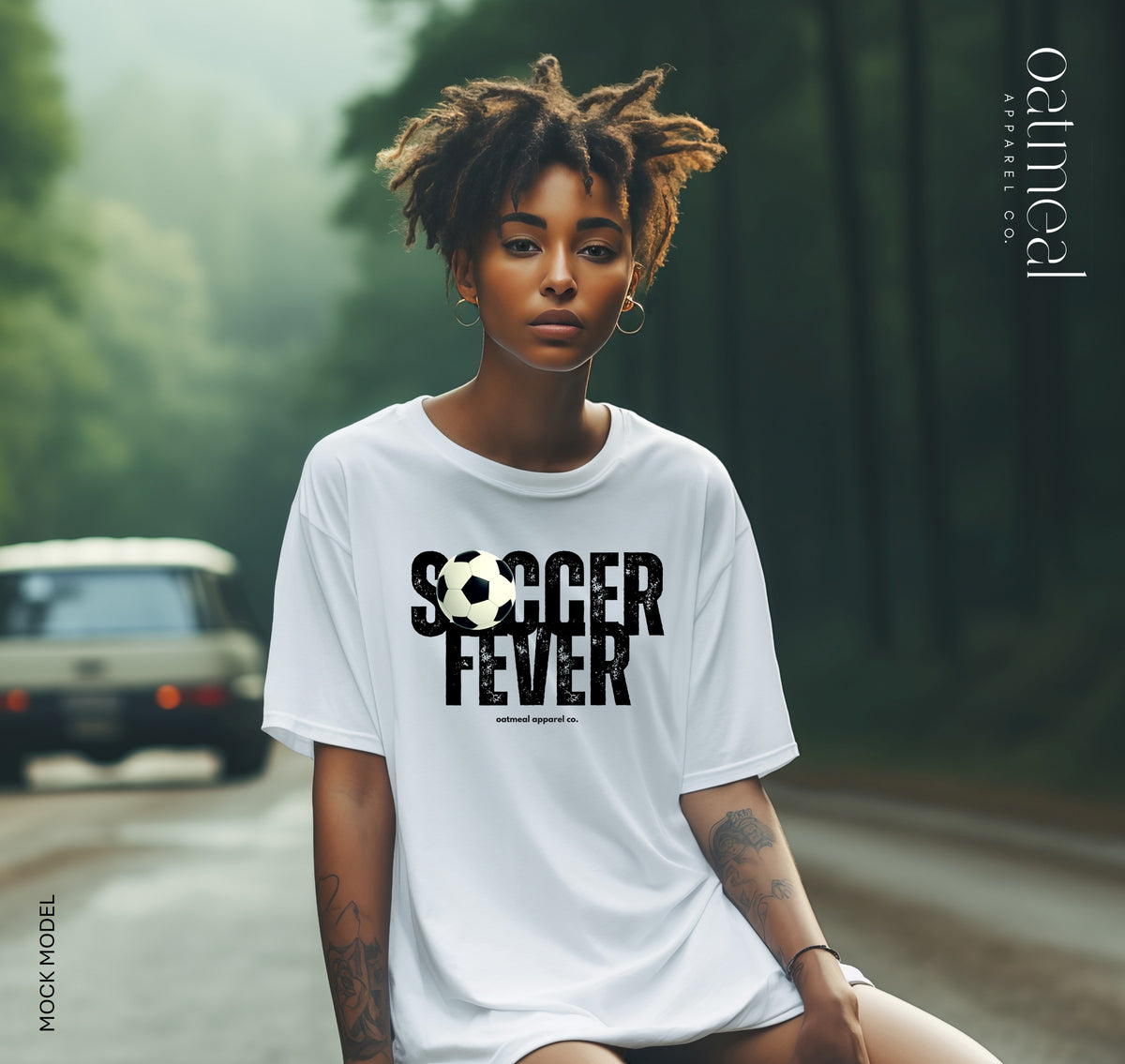 A woman wearing a white graphic t-shirt with 'SOCCER FEVER' and a soccer ball graphic printed on the front, set against a blurred forest road background with a vintage car model for Oatmeal Apparel Co.