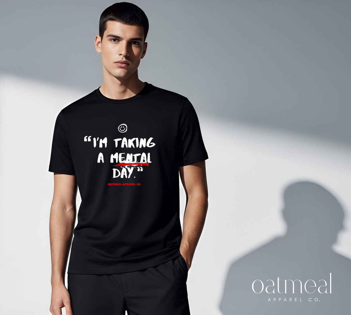 A male model wearing a black graphic t-shirt with the text 'I'M TAKING A MENTAL DAY.' in white, set against a gray background with a soft shadow at Oatmeal Apparel Co.