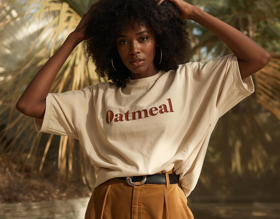 A woman with an afro hairstyle wearing a beige graphic t-shirt with the word 'Oatmeal' printed in dark red across the chest, paired with brown pants and a black belt, posing outdoors with palm trees in the background for Oatmeal Apparel Co.