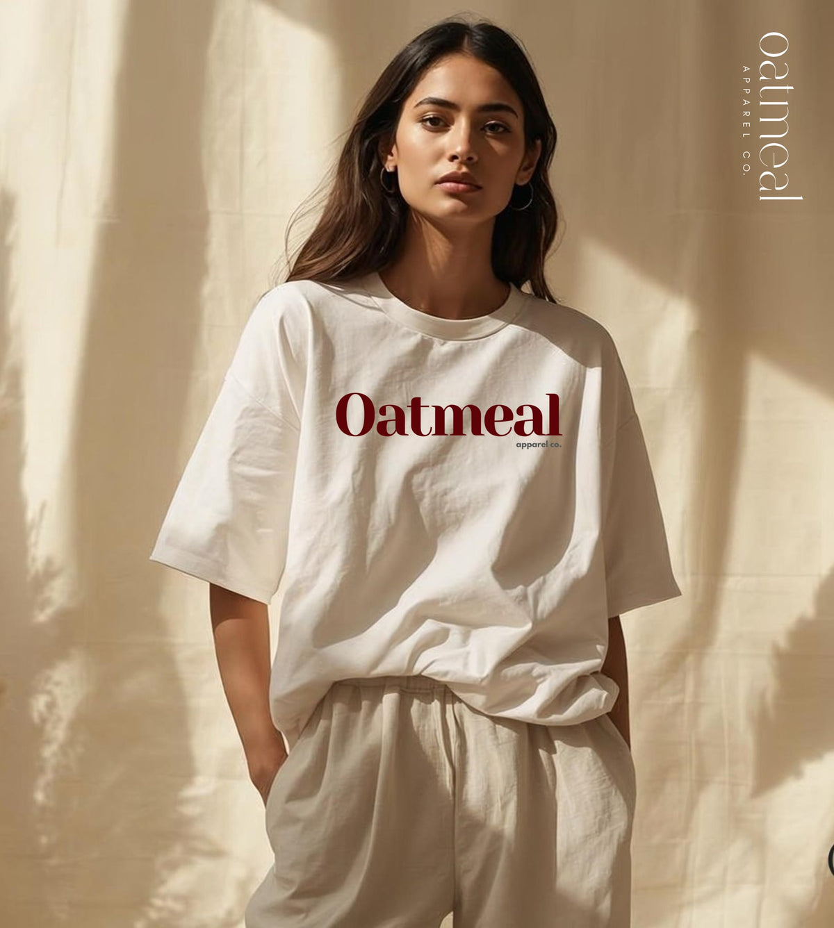 A woman wearing a cream-colored oversized organic cotton T-shirt with 'Oatmeal' printed in dark red across the chest, paired with matching cream pants, standing against a draped beige fabric backdrop with soft shadows at Oatmeal Apparel Co.