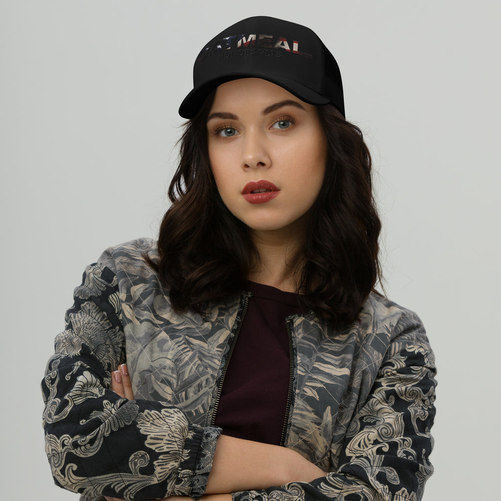 Woman wearing 'honors vets' black cap and patterned jacket against a plain background at Oatmeal Apparel Co.