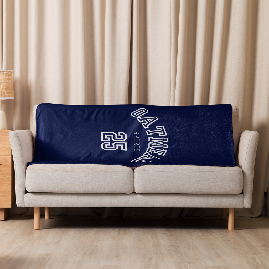 Navy blue blanket with 'oatmeal sports 25' text on a beige sofa against a beige curtain background at Oatmeal Apparel Co.
