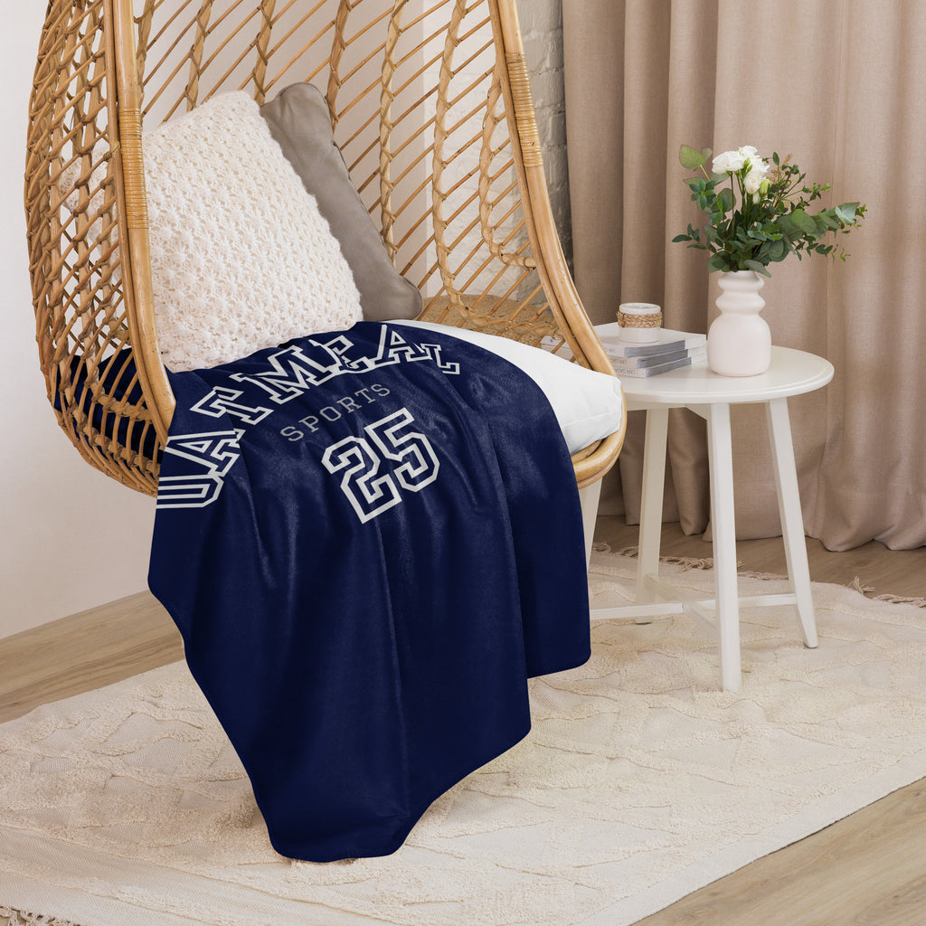 Navy blue sports-themed blanket with white text draped over a wicker chair in a cozy room at Oatmeal Apparel Co.