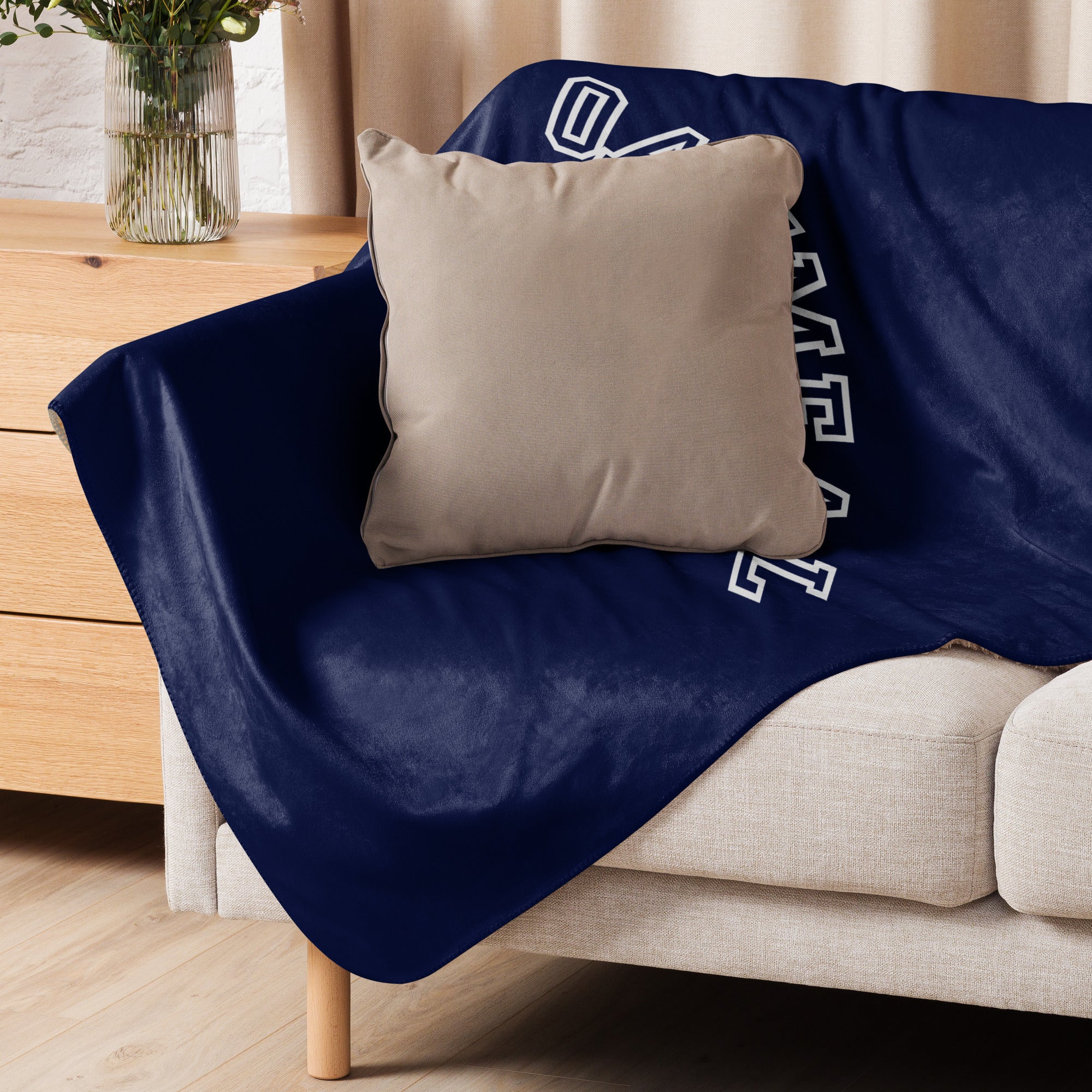 Navy blanket with sports-themed design draped over a beige sofa with a beige pillow at Oatmeal Apparel Co.