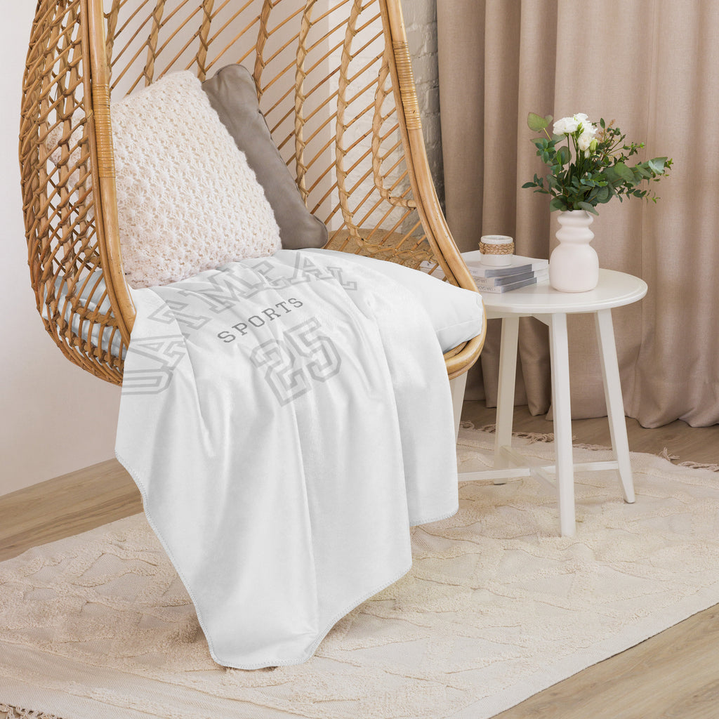 White blanket with 'Oatmeal Sports 25' text draped over a wicker chair in a cozy room, exclusively at Oatmeal Apparel Co.