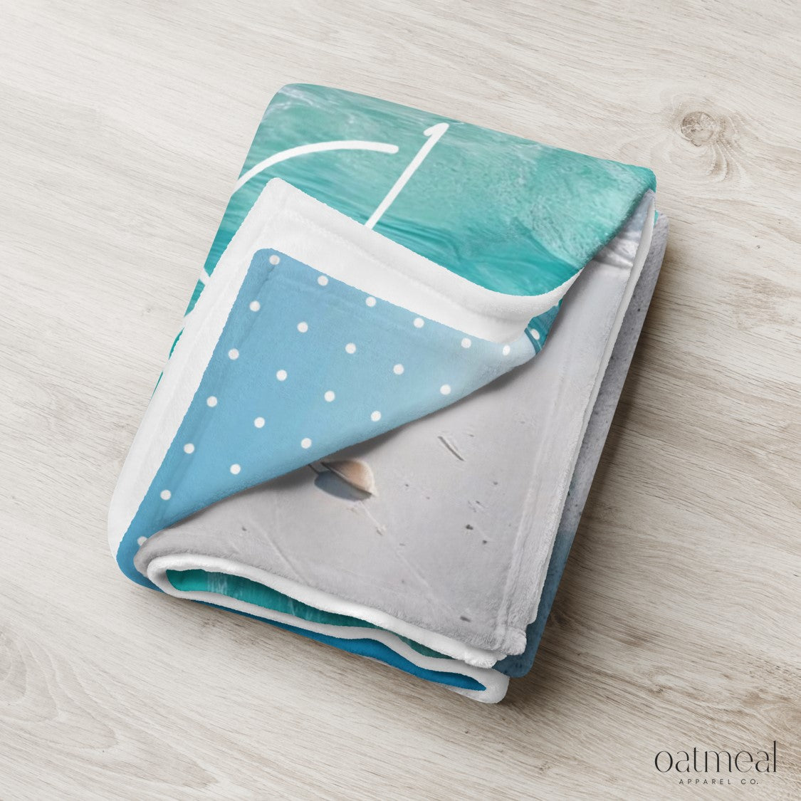 Folded blanket with teal, white, and gray pattern on a light wooden surface at Oatmeal Apparel Co.