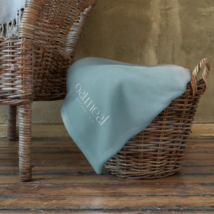 Light blue Wolfdog design on front of blanket with 'Oatmeal' branding draped over a wicker basket on a wooden floor at Oatmeal Apparel Co.