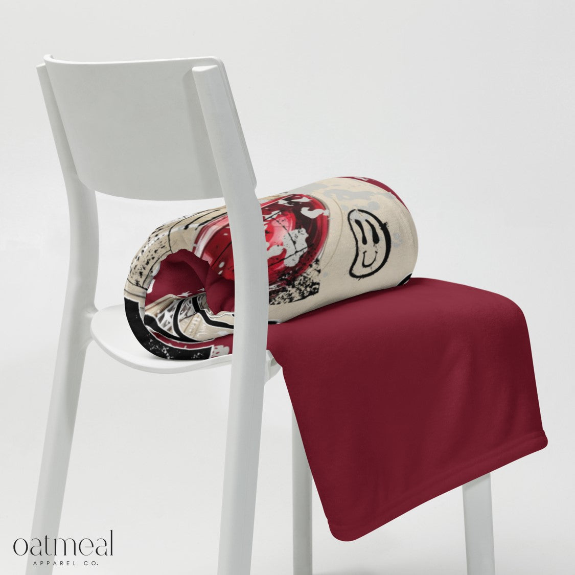 White chair with folded dark red throw blanket and a patterned roll, featuring the Oatmeal Apparel Co. logo.