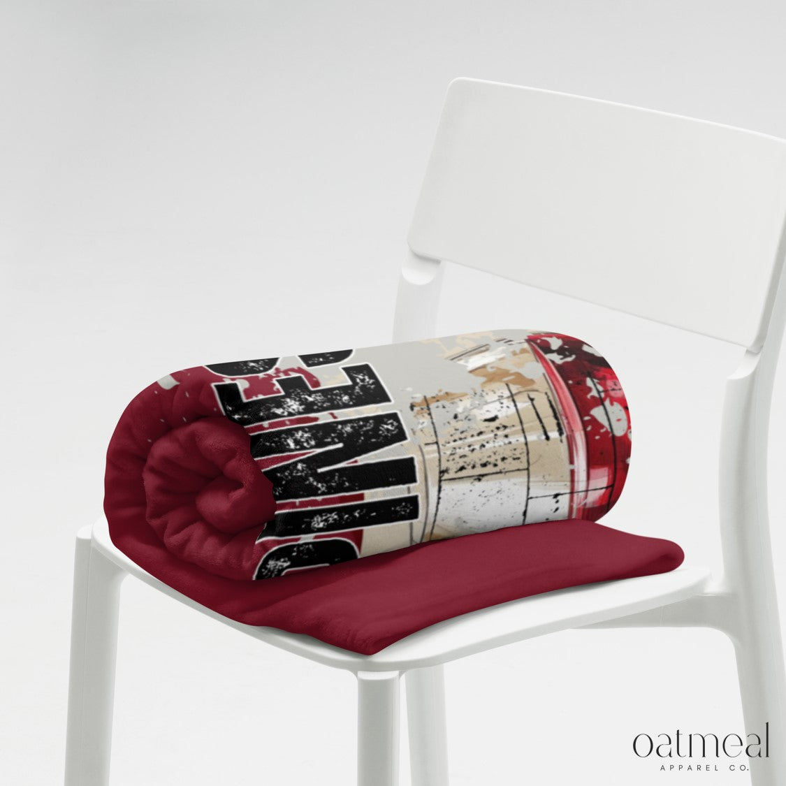 Dark red cylindrical style folded blanket with a red base and patterned top on a white chair, brand 'oatmeal' visible