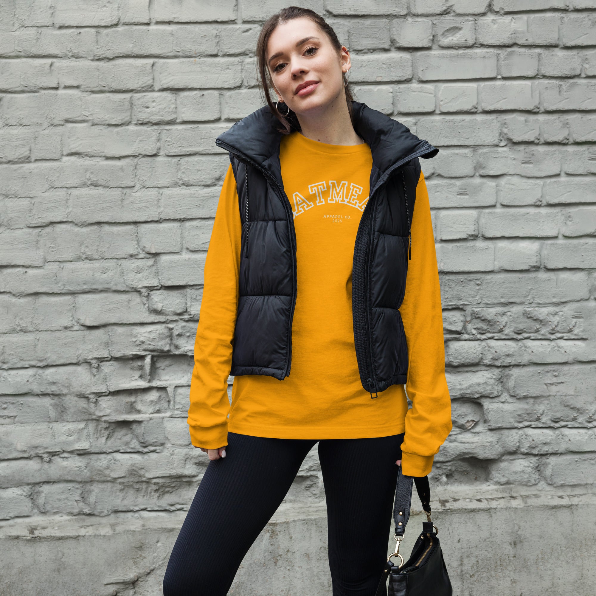 Woman wearing a gold long-sleeve shirt with a black puffer vest against a brick wall at Oatmeal Apparel Co., Sports Edition Design.