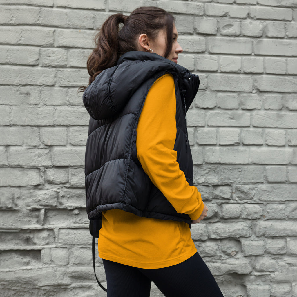 Person wearing a black puffer vest over a yellow long-sleeve shirt against a brick wall, shirt avail at Oatmeal Apparel Co., Sports Edition Design.
