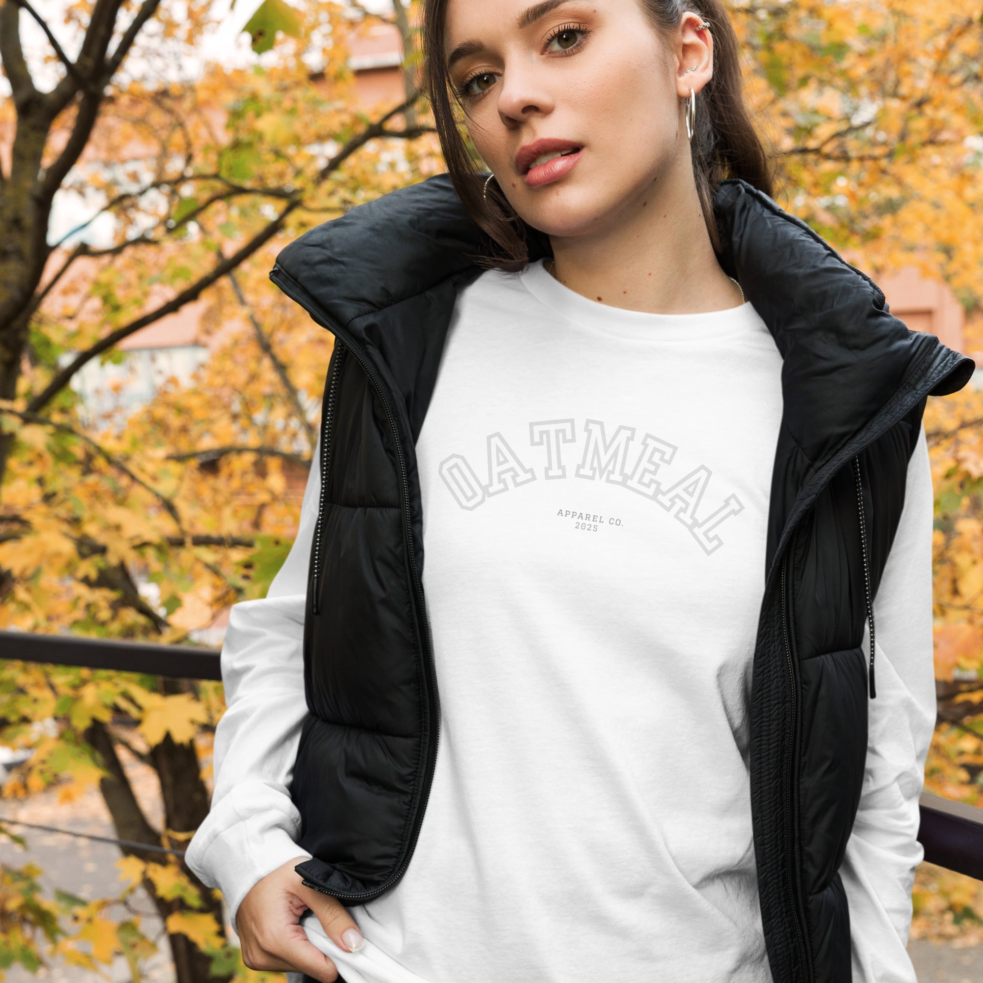Woman wearing a white 'OATMEAL' long-sleeve shirt with a black puffer vest in an autumn setting at Oatmeal Apparel Co., Sports Edition Design.