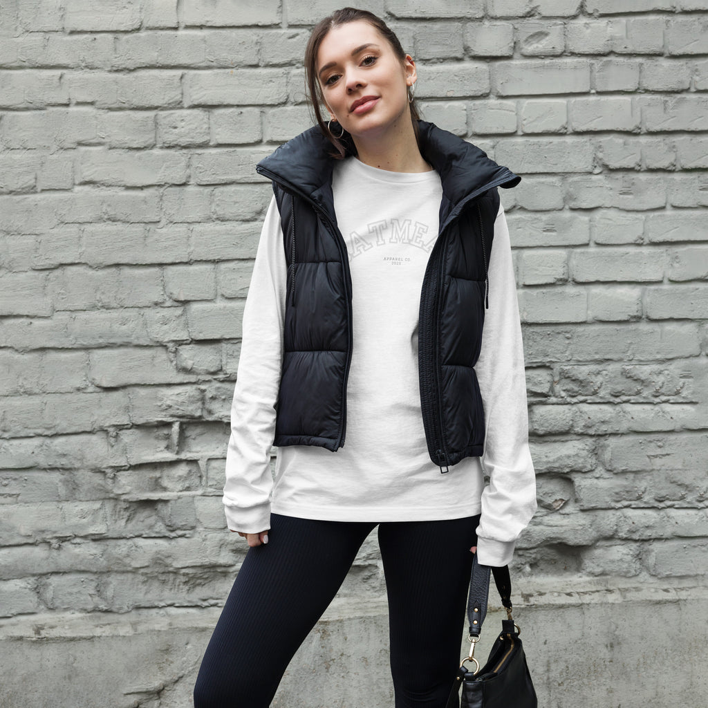 Woman wearing a white long sleeve shirt, black puffer vest, and black leggings against a brick wall at Oatmeal Apparel Co., Sports Edition Design.