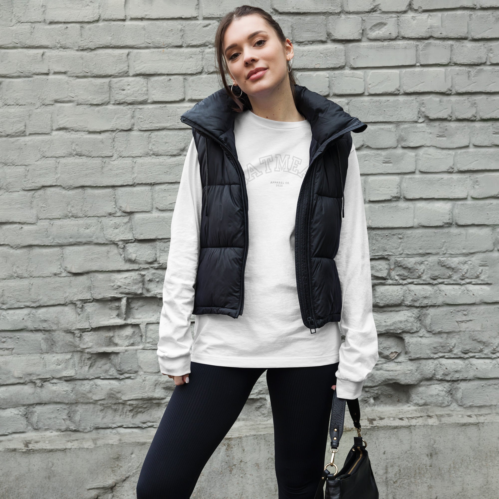 Woman wearing a white long sleeve shirt, black puffer vest, and black leggings against a brick wall at Oatmeal Apparel Co., Sports Edition Design.