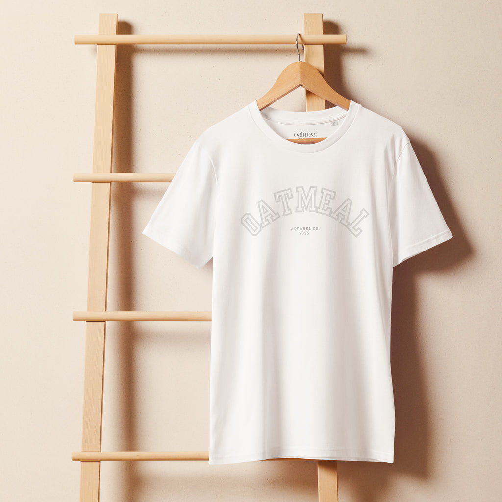 White t-shirt with 'Oatmeal' sports branding on a wooden hanger against a beige wall at Oatmeal Apparel Co.