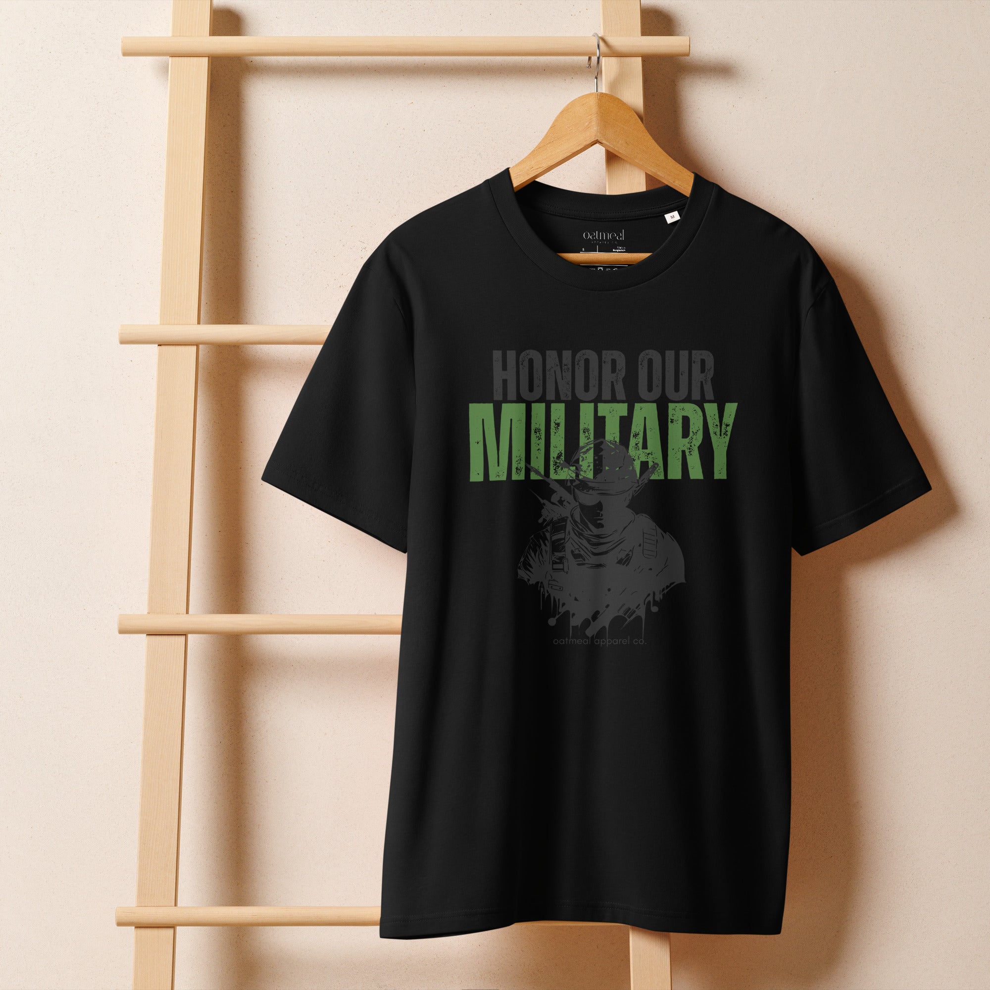 Black t-shirt with 'Honor Our Military' text on a wooden hanger against a beige wall available at Oatmeal Apparel Co.