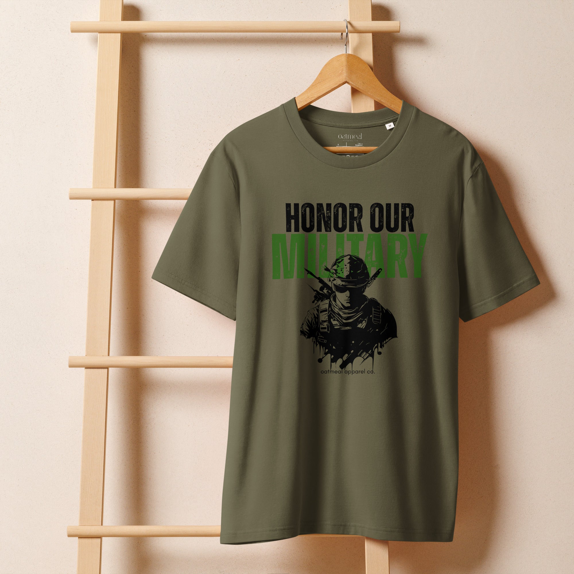 Green t-shirt with 'Honor Our Military' text on a wooden hanger against a beige wall avail at Oatmeal Apparel Co.