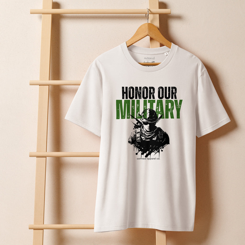 White t-shirt with 'Honor Our Military' text and graphic on a wooden hanger against a beige background at Oatmeal Apparel Co.