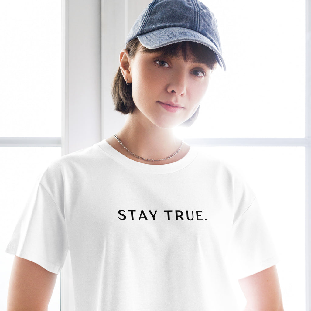 Person wearing a white crop top with 'STAY TRUE.' text and a blue cap indoors, available at Oatmeal Apparel Co.