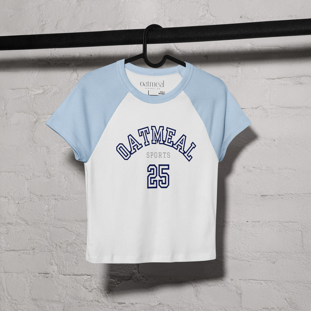 White and light blue baseball-style t-shirt with 'Oatmeal Sports' branding on a hanger against a gray brick wall.