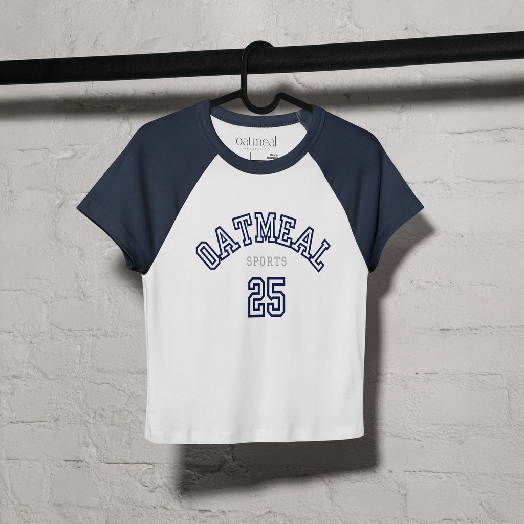 White and navy baseball shirt with 'Oatmeal Sports' text on a hanger against a light gray brick wall at Oatmeal Apparel Co.