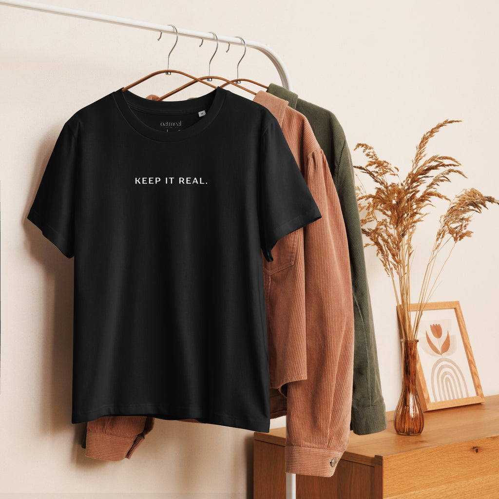 Women's Organic Cotton Black t-shirt with 'KEEP IT REAL.' text on a hanger with other clothes in the background at Oatmeal Apparel Co.